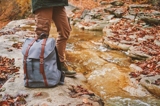 Unrecognizable female hiker with backpack standing near a river in autumn forest, view of legs