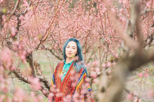 Beautiful boho style young woman walking in blooming peach tree garden, looking at camera.