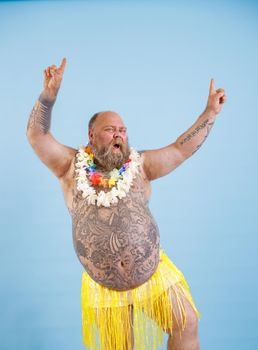 Jouful bearded plump man with bare chest in decorative yellow grass skirt and flowers garlands has fun on light blue background in studio