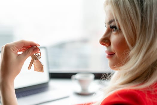 Mortgage concept. Woman in red business suit holding key with house shaped keychain. Modern light lobby interior. Real estate, moving home or renting property