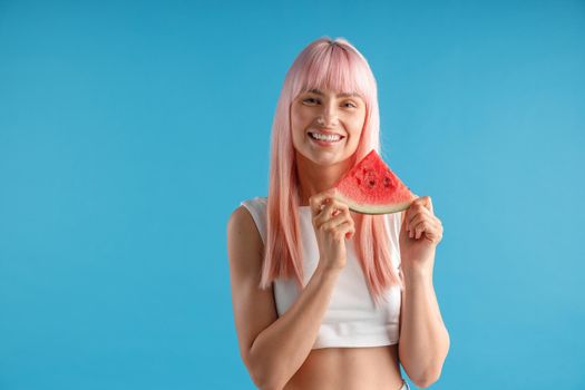 Smiling young woman with natural long pink dyed hair holding a slice of watermelon, posing isolated over blue studio background. Beauty and health concept