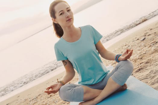 Young woman meditating in pose of lotus on sand shore on background of sea, looking at camera.