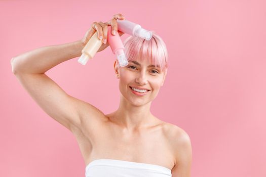 Pleased young woman with pink hair holding three plastic travel bottles with beauty products, posing isolated over pink background. Beauty, spa, body care concept