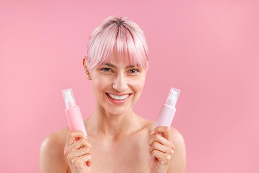 Portrait of gorgeous woman with pink hair smiling and holding two plastic bottles with beauty products, posing isolated over pink background. Beauty, spa, body care concept