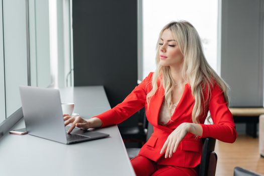 Woman in red coral business suit. Young blonde businesswoman with modern gray laptop in the office in high floor in front of the window. Girl drinking coffee.