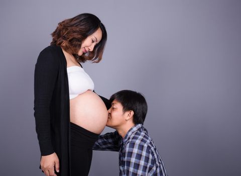 Young man kissing his pregnant wife's belly on gray wall background