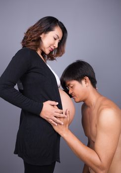 Young man kissing his pregnant wife's belly on gray wall background