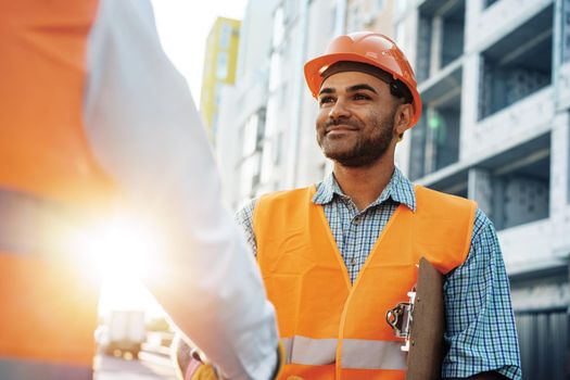 Two men engineers in workwear shaking hands against construction site, close up