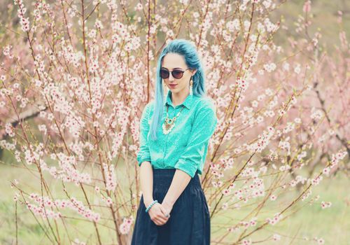 Attractive fashionable young woman with blue hair walking in blossoming peach tree garden in spring.