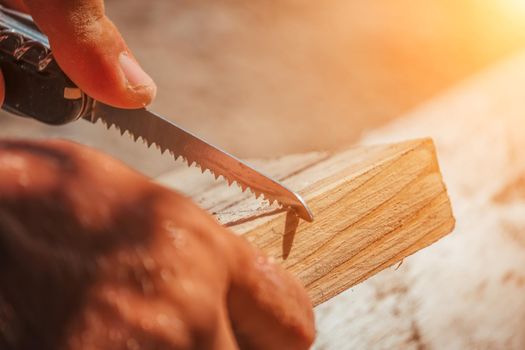 Man Hands using a Swiss knife, sawing a piece of wood plank, outdoor survival and camping, fun in the woods, nobody.