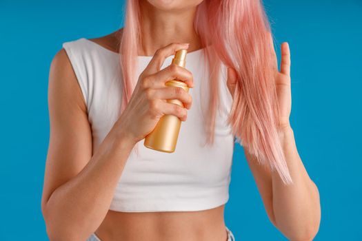 Close up shot of hands of young woman holding and applying moisturizing spray to her smooth pink hair, standing isolated over blue studio background. Beauty products, hair care concept