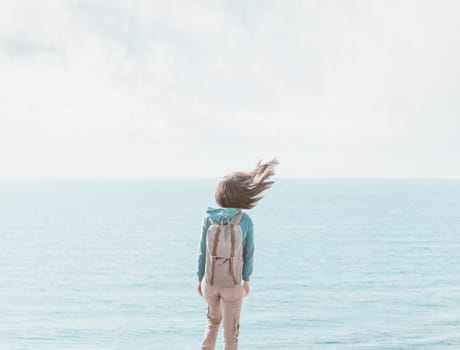 Traveler young woman with backpack standing on background of sea in windy weather, her hair fluttering in the wind, rear view.