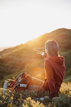 Backpacker young woman resting in summer mountains at sunset. Female hiker drinking tea from thermos.