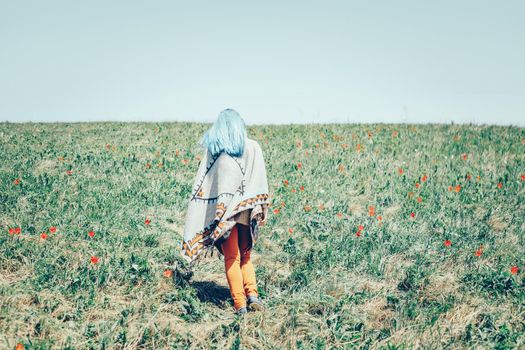 Unrecognizable young woman with blue hair wearing in poncho walking in wildflower field.