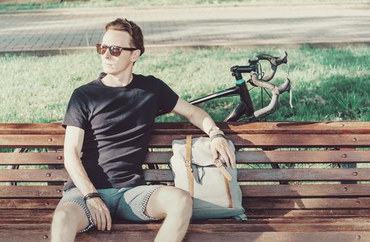 Handsome guy cyclist sitting with backpack on wooden bench in summer park.