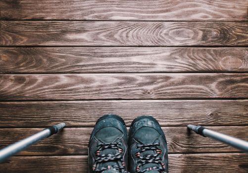 Hiking boots with trekking poles on wooden background, point of view. Copy-space.