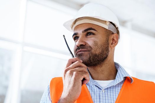 Young construction worker in uniform using walkie talkie on site, close up portrait