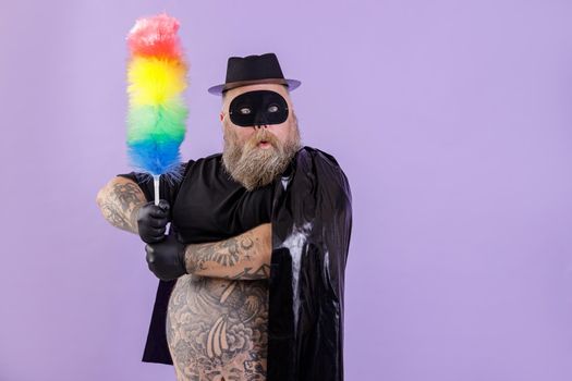 Stylish middle aged plump man in Zorro suit with tattoos on bare belly holds colorful dust brush on purple background in studio