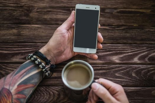 Young man sitting with a smartphone and a cup of coffee at a wooden table, point of view. Free space on screen of phone.