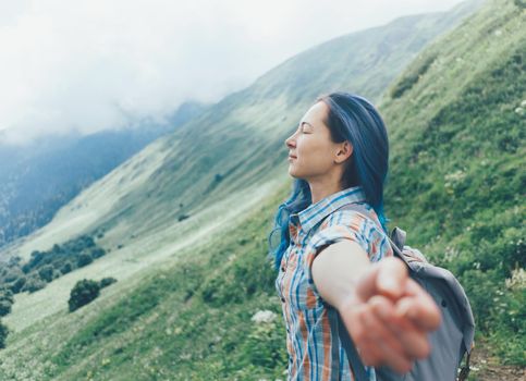 Freedom explorer young woman with backpack standing with raised arms in summer mountains, side view.