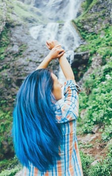 Young woman with blue hair standing with raised arms in front of waterfall in summer.