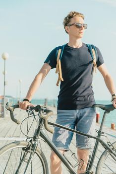 Handsome young man in sunglasses standing with bicycle on pier in summer, close-up.