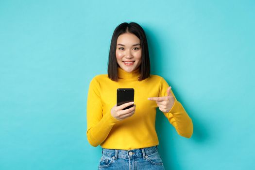 E-commerce and online shopping concept. Cute asian woman in yellow sweater pointing at smartphone, smiling at camera, standing over blue background.