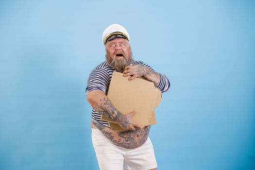 Happy bearded obese man in sailor costume holds blank beige boxes of pizza posing on light blue background in studio