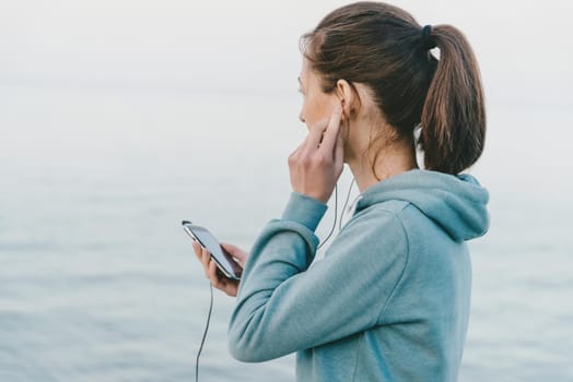 Fitness young woman listens music in headphones on her smartphone for workout on background of sea.