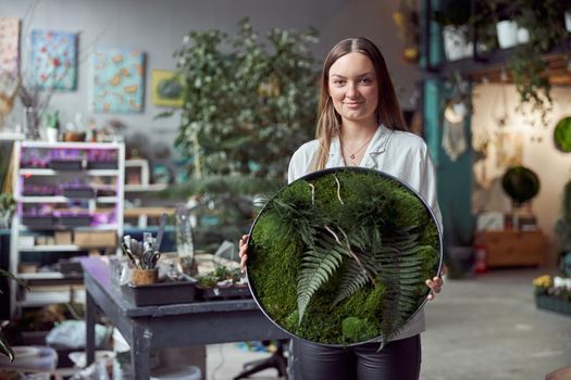 Confident female florist is holding a plant composition in her own flower and plants shop