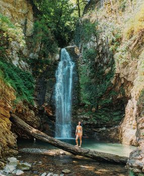 Beautiful young woman standing on fallen tree trunk in front of waterfall in summer.