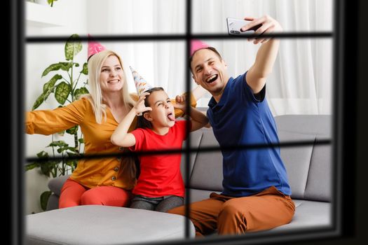 Young big family celebrating Christmas enjoying dinner, view from outside through a window into a decorated living room with tree and candle lights, happy parents eating with three kids