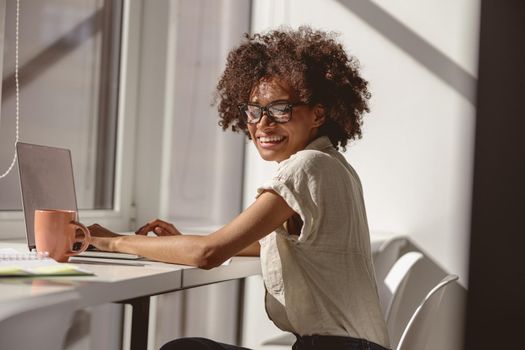 Happy Afro American lady in glasses enjoying her job in the office while typing on laptop