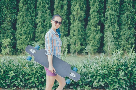 Beautiful girl in sunglasses walking with longboard in summer park.