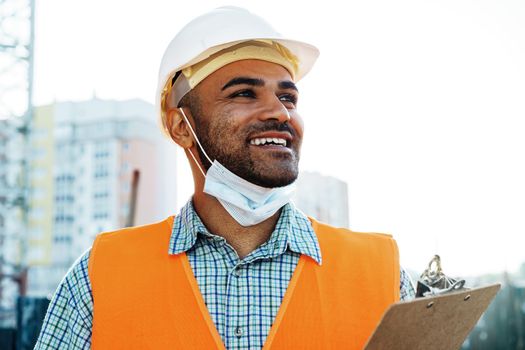 Portrait of mixed race man builder in workwear and hardhat wearing medical mask, close up photo