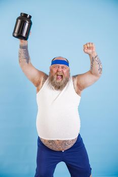 Positive mature bearded man with overweight raises up hand and large bottle of sport nutritient standing on light blue background in studio