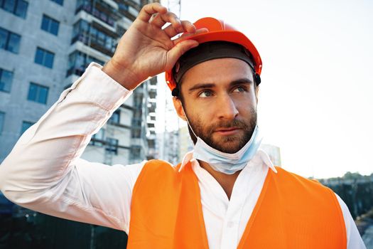 Portrait of man builder in workwear and hardhat wearing medical mask, close up photo