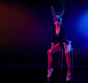 the beautiful girl in a bodysuit poses in a photo Studio on a dark background in neon light. On the face of a rabbit mask