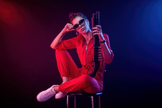 bright brunette in a red jumpsuit with a shotgun on a dark background in neon light