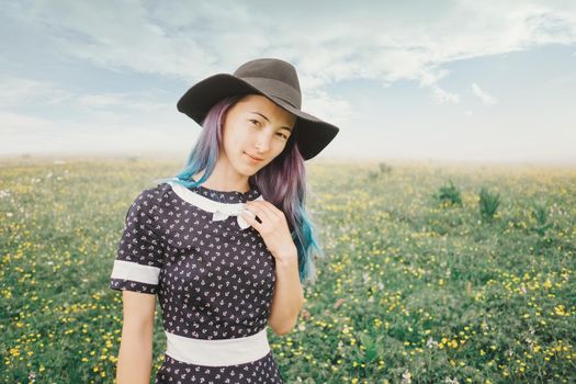 Beautiful young woman wearing in dress and hat walking on wildflower meadow, looking at camera.