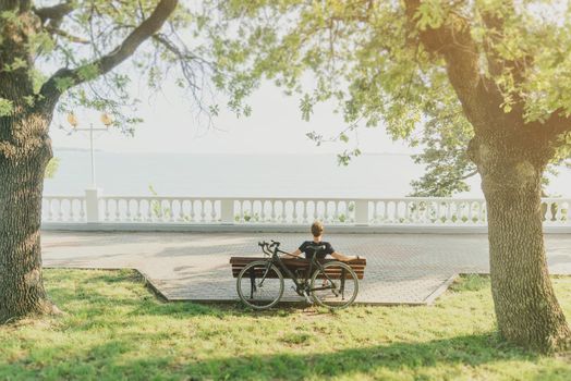 Young man rests with bicycle on wooden bench in summer park on coastline, rear view.