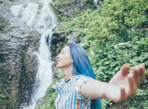 Happy young woman standing with raised arms on background of waterfall and enjoying of summer nature.
