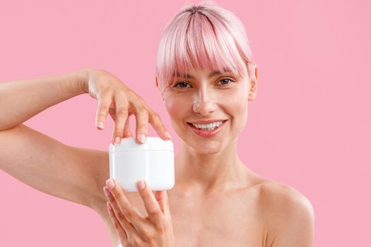 Portrait of lovely young woman with pink hair holding white jar with moisturizing body lotion after shower, posing isolated over pink background. Beauty, spa, body care concept