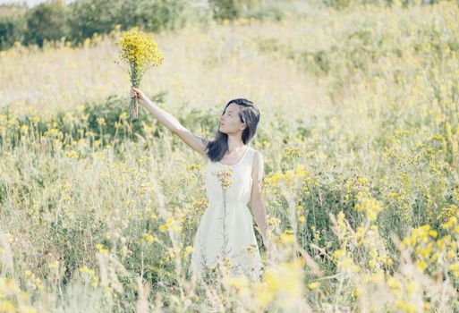 Beautiful young woman with bouquet of flowers walking on summer meadow.