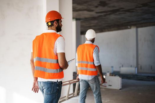 Two young male engineers in uniform and hardhats working at construction site, close up