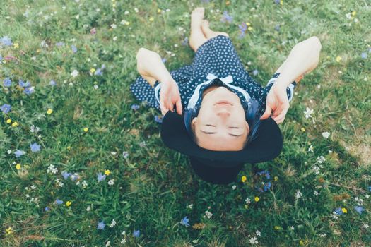 Beautiful young woman wearing in dress and hat resting on wildflower summer meadow.