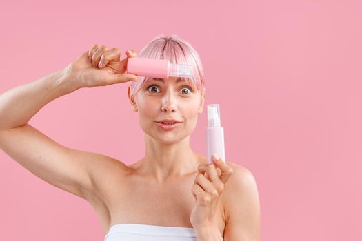 Portrait of cute woman with pink hair looking surprised, holding two plastic bottles with beauty products, posing isolated over pink background. Beauty, spa, body care concept