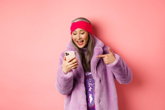 Online shopping and fashion concept. Cool asian senior woman checking out promotion on mobile phone, pointing finger at smartphone screen and smiling pleased, pink background.