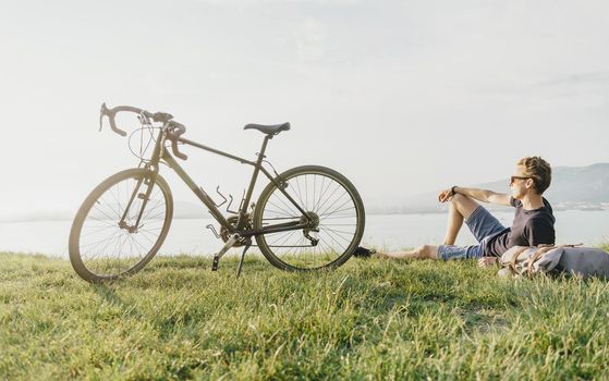 Young man cyclist rests on summer meadow near the bicycle.