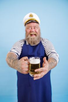 Smiling middle aged obese man in sailor costume with apron stands on light blue background in studio, focus on hands with mug of beer